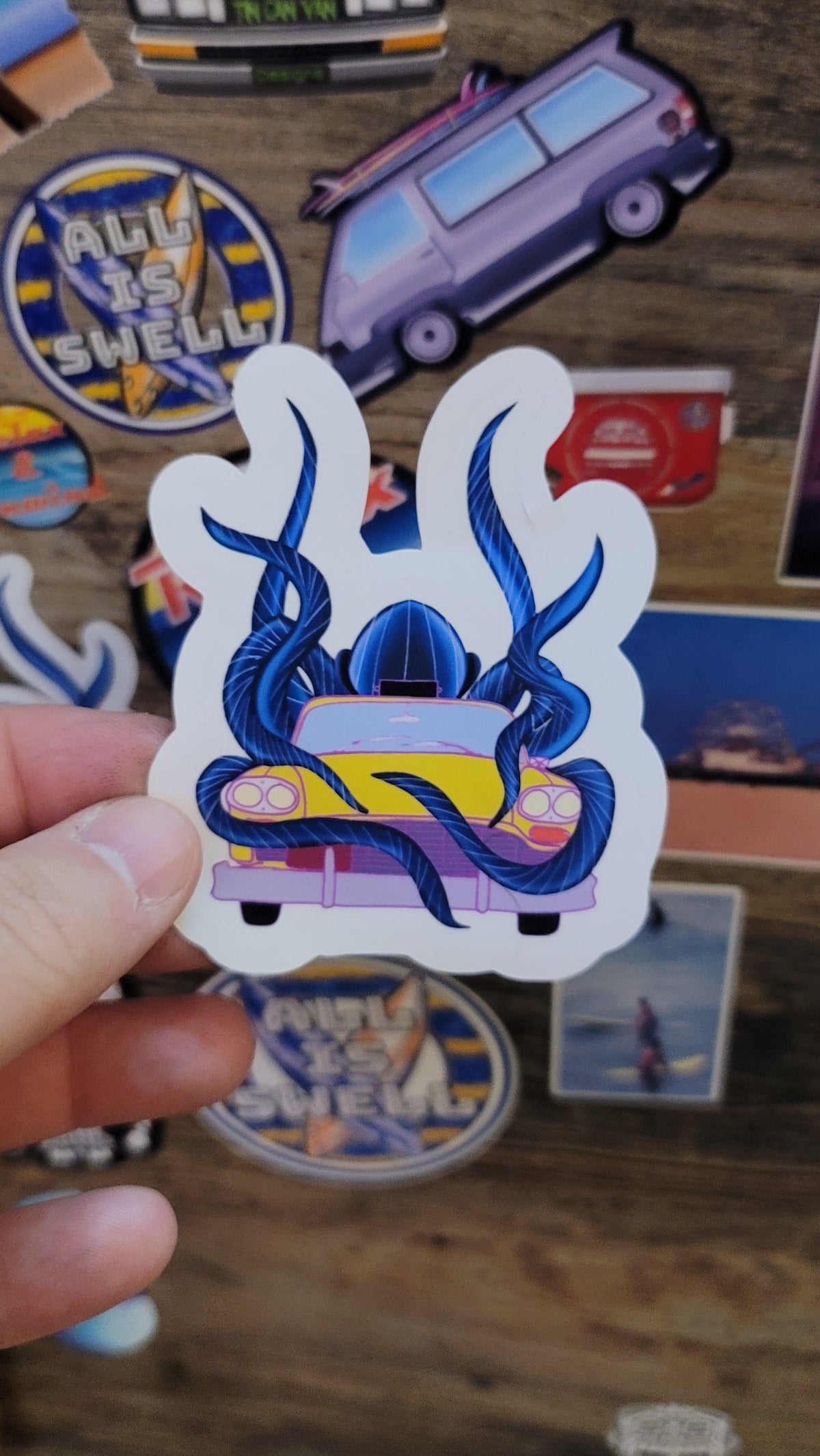 Hand holding a colorful sticker of an octopus engulfing a yellow taxi cab against a wooden background with other stickers.