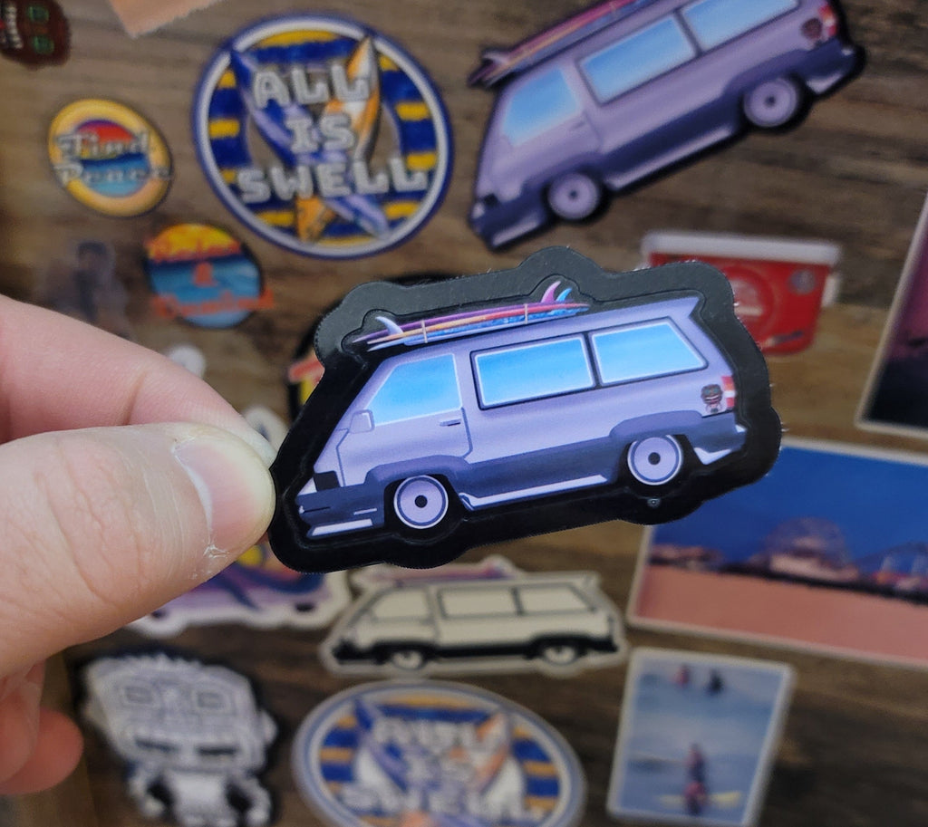 Hand holding a sticker of a surf van among other colorful stickers on a wooden surface