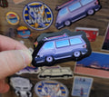 Hand holding a sticker of a surf van among other colorful stickers on a wooden surface
