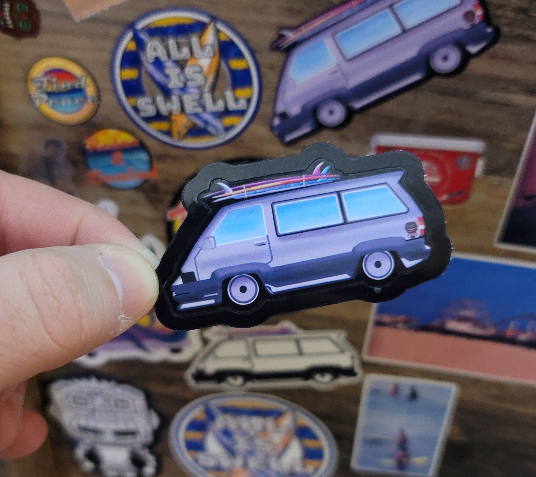 Hand holding a sticker of a surf van among other colorful stickers on a wooden surface
