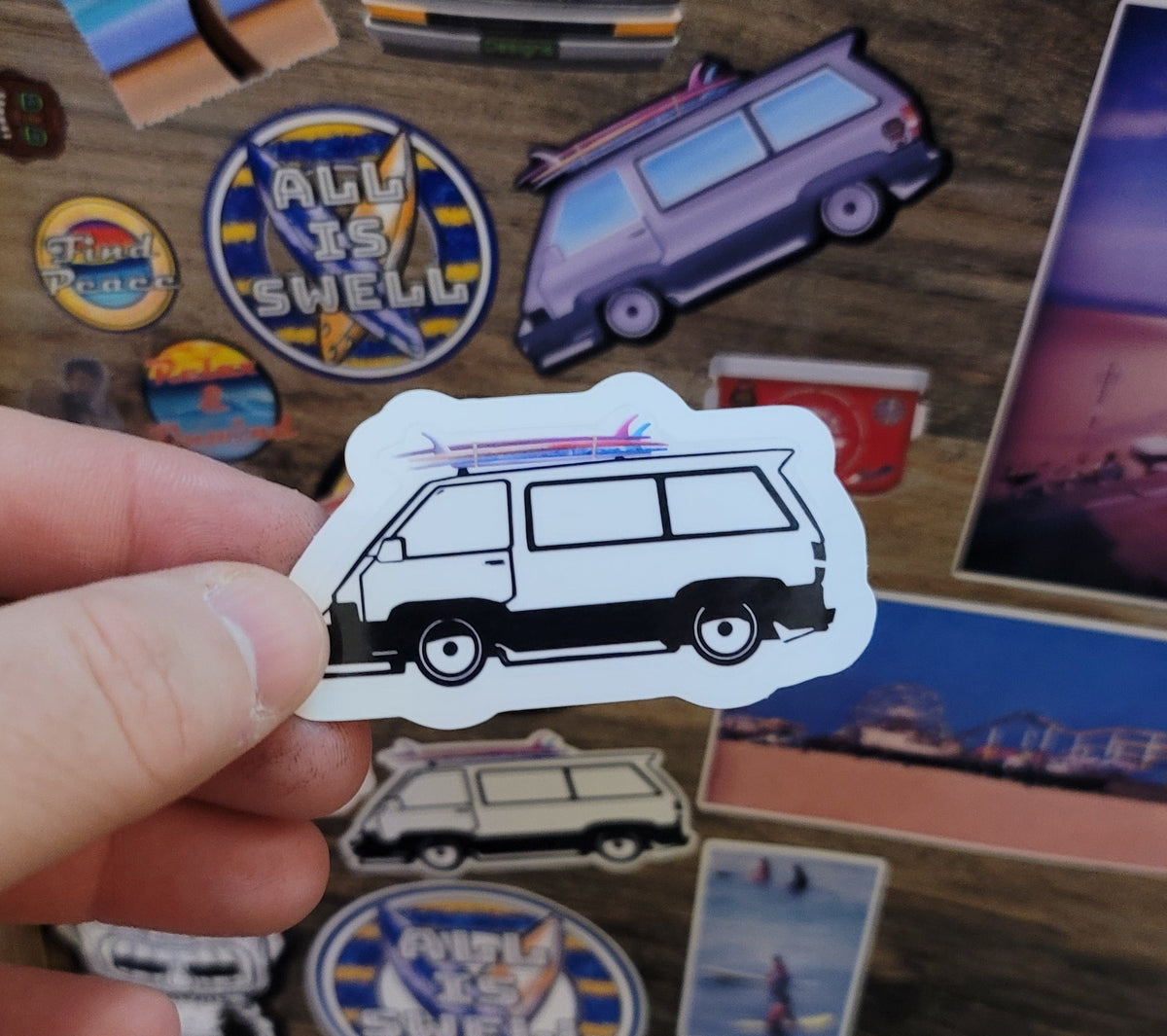 Hand holding a sticker of a surf van among other colorful stickers on a wooden surface