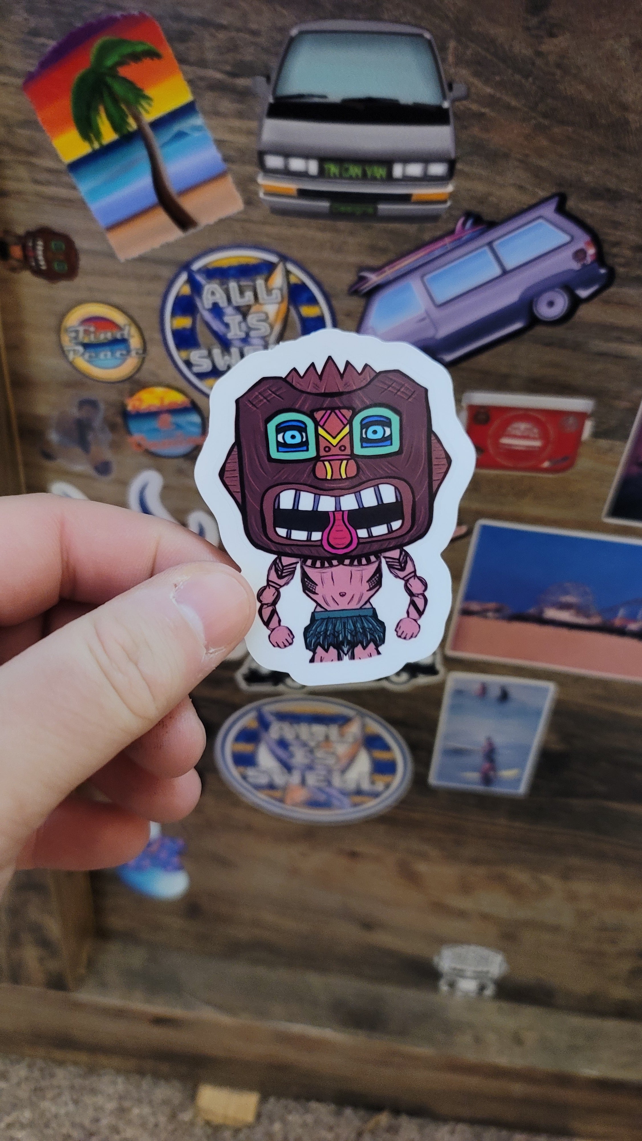 Hand holding a colorful tiki guy sticker with a cartoonish design against a background of other stickers.