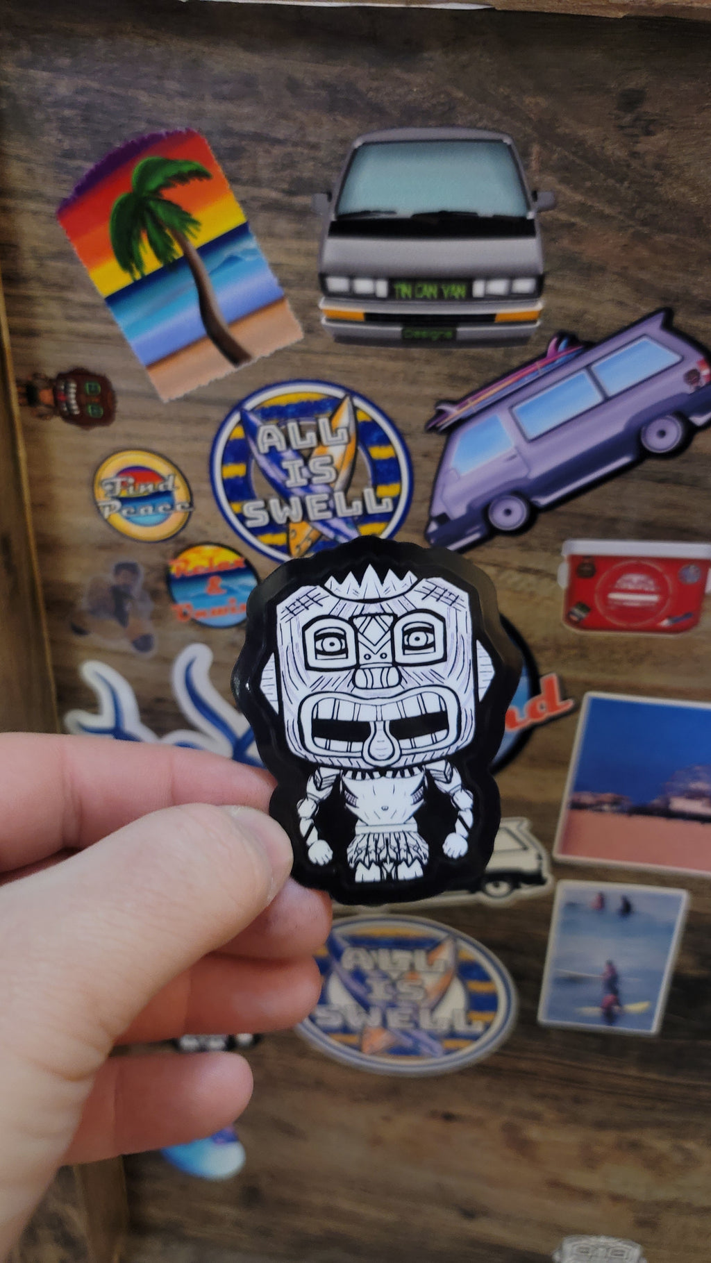 Hand holding a black and white tiki guy sticker over a collection of colorful stickers on a wooden surface