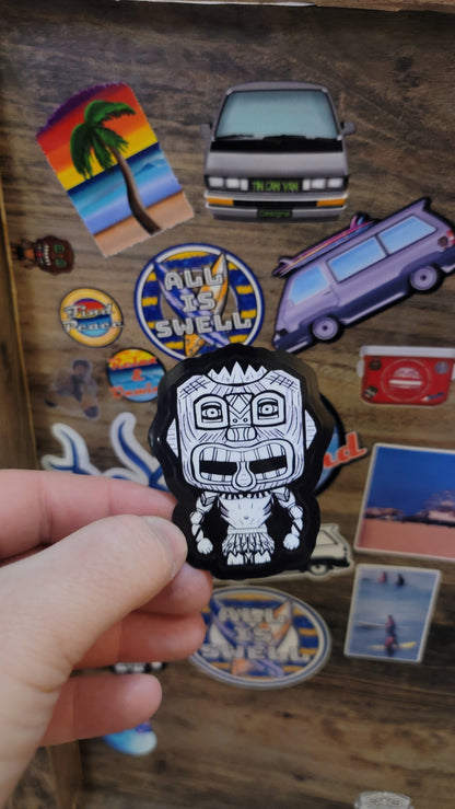 Hand holding a black and white tiki guy sticker over a collection of colorful stickers on a wooden surface
