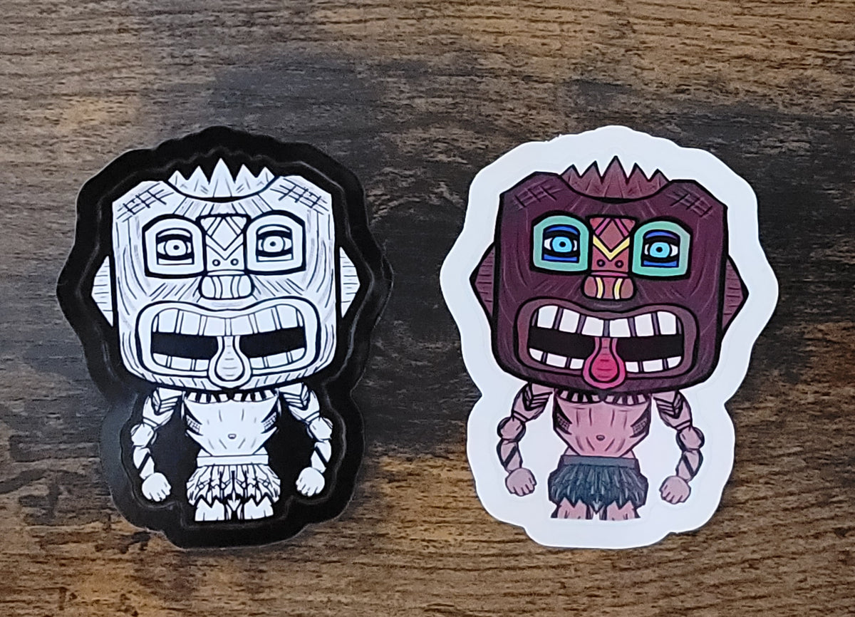 Two tiki guy stickers, one black and whitecand the other full color, on a wooden surface