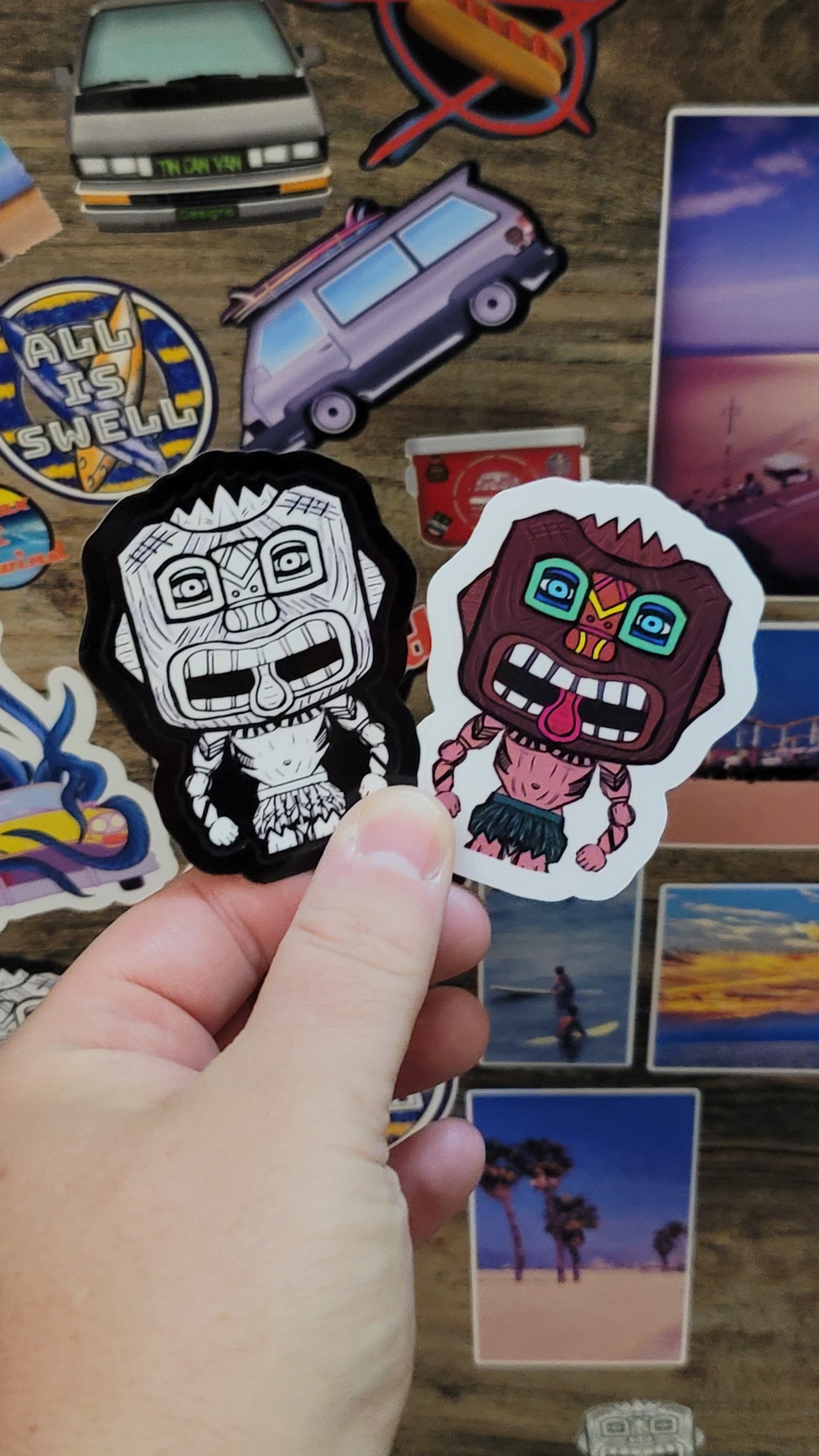 Hand holding two tiki guy stickers, one black and whitecand the other full color, against a wooden surface with other stickers.