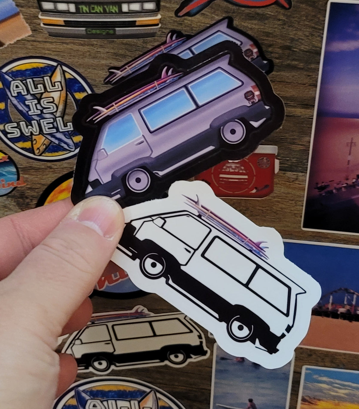 Hand holding 2 stickers , one black and white and one full color, of a surf van among other colorful stickers on a wooden surface