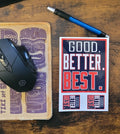 Mouse, mouse pad with tiki design, pen, and 'Good. Better. Best.' Sticker sheet on a wooden surface