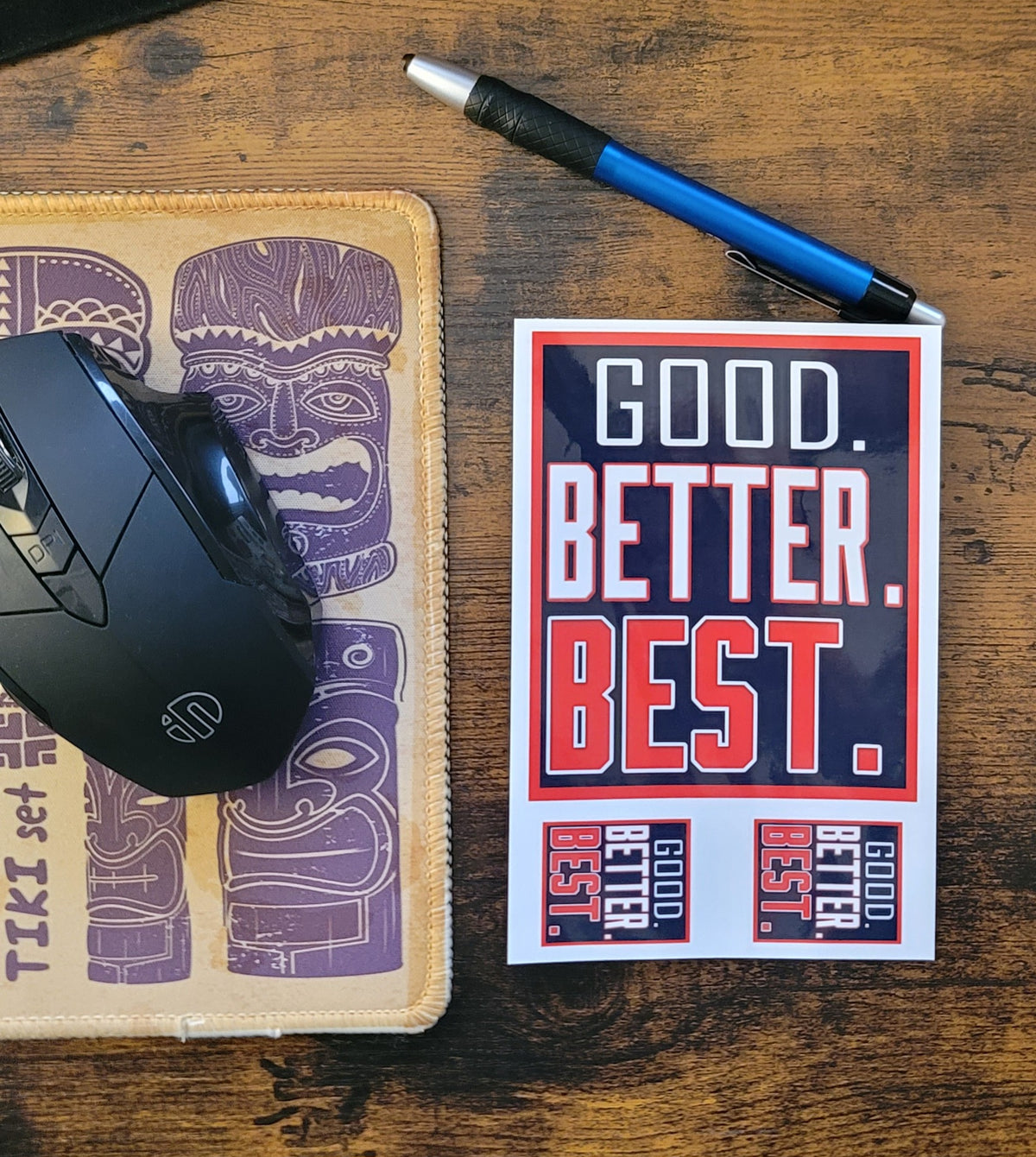 Mouse, mouse pad with tiki design, pen, and 'Good. Better. Best.' Sticker sheet on a wooden surface