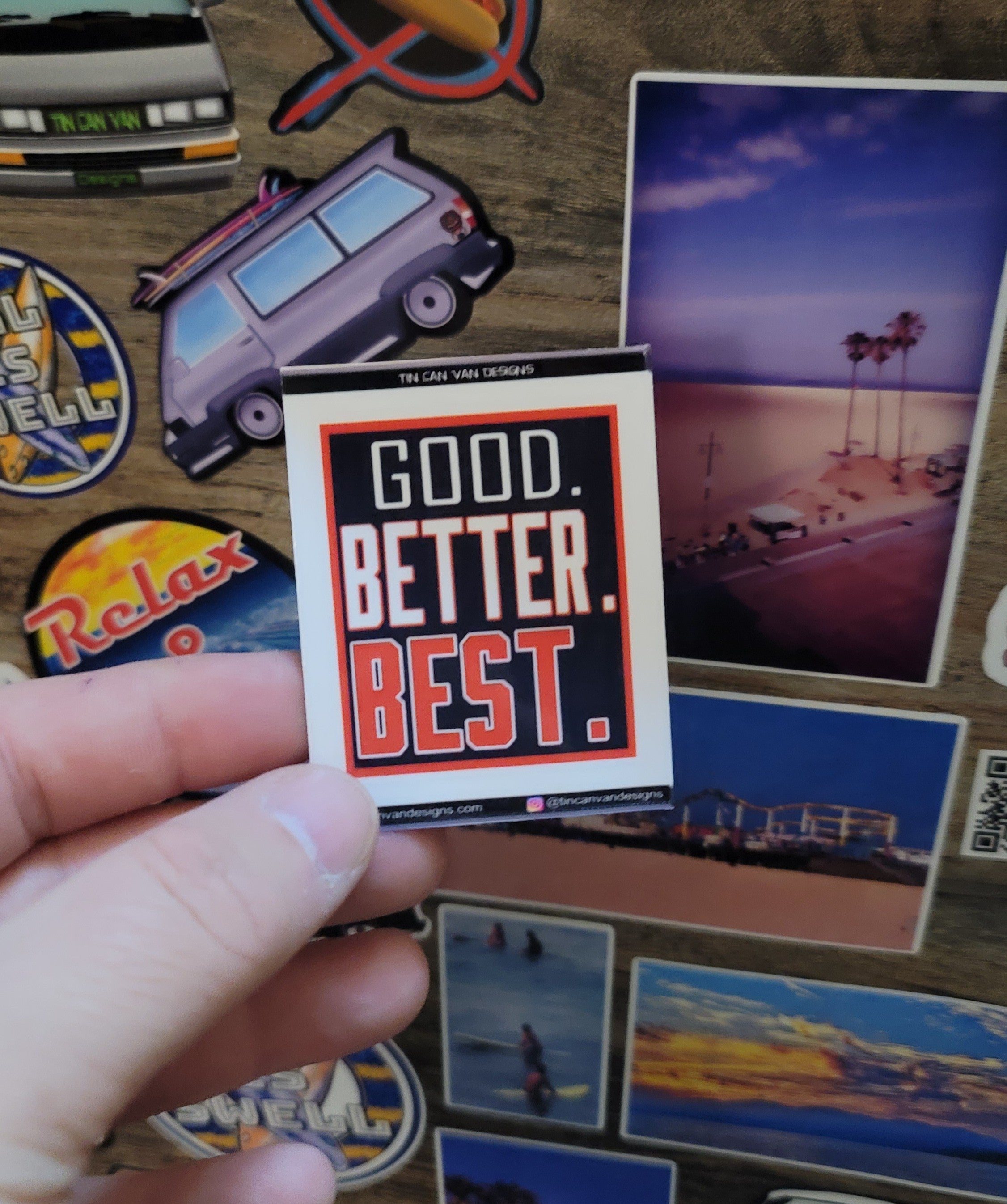 1.75" x 2" Sticker with 'Good. Better. Best.' text held over a collection of other stickers on a textured surface.