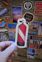 Hand holding a red and white dive flag scuba diving tank sticker against a background of various other stickers.