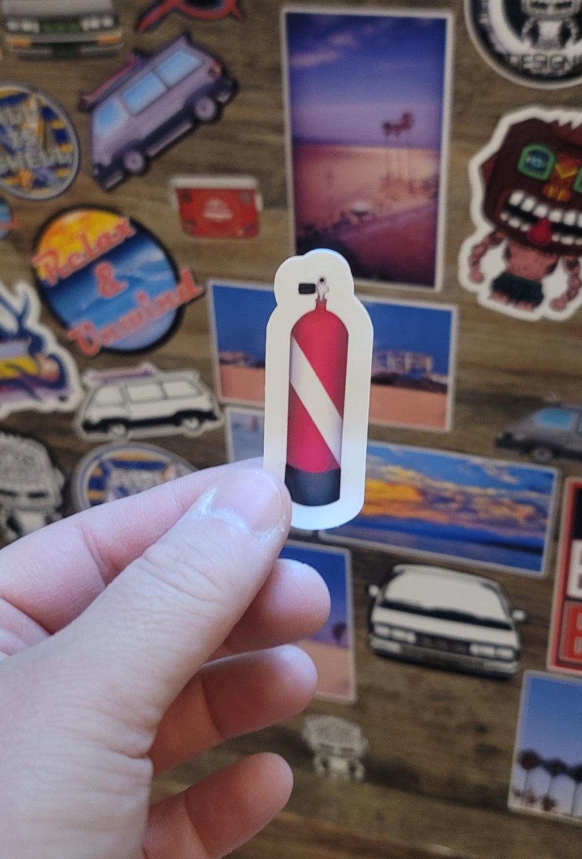 Hand holding a red and white dive flag scuba diving tank sticker against a background of various other stickers.