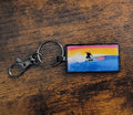 Keychain with a drawing of a surfer riding a wave against a sunset background on a wooden surface.