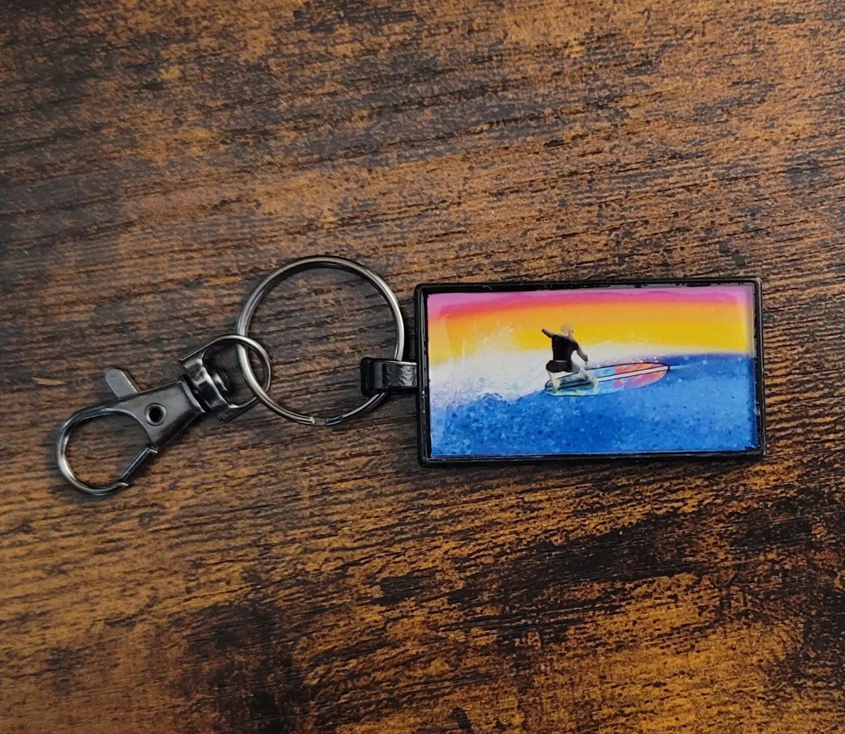 Keychain with a drawing of a surfer riding a wave against a sunset background on a wooden surface.