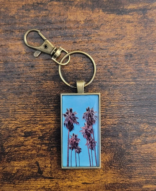 Gold keychain of a photo of a group of palm trees with a blue/aqua sky background on top of a wooden surface.