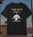 A black short-sleeve t-shirt with a white graphic print of a palm tree and the text 'LONE PALM SOCIAL CLUB' on the back, displayed against a beach background.