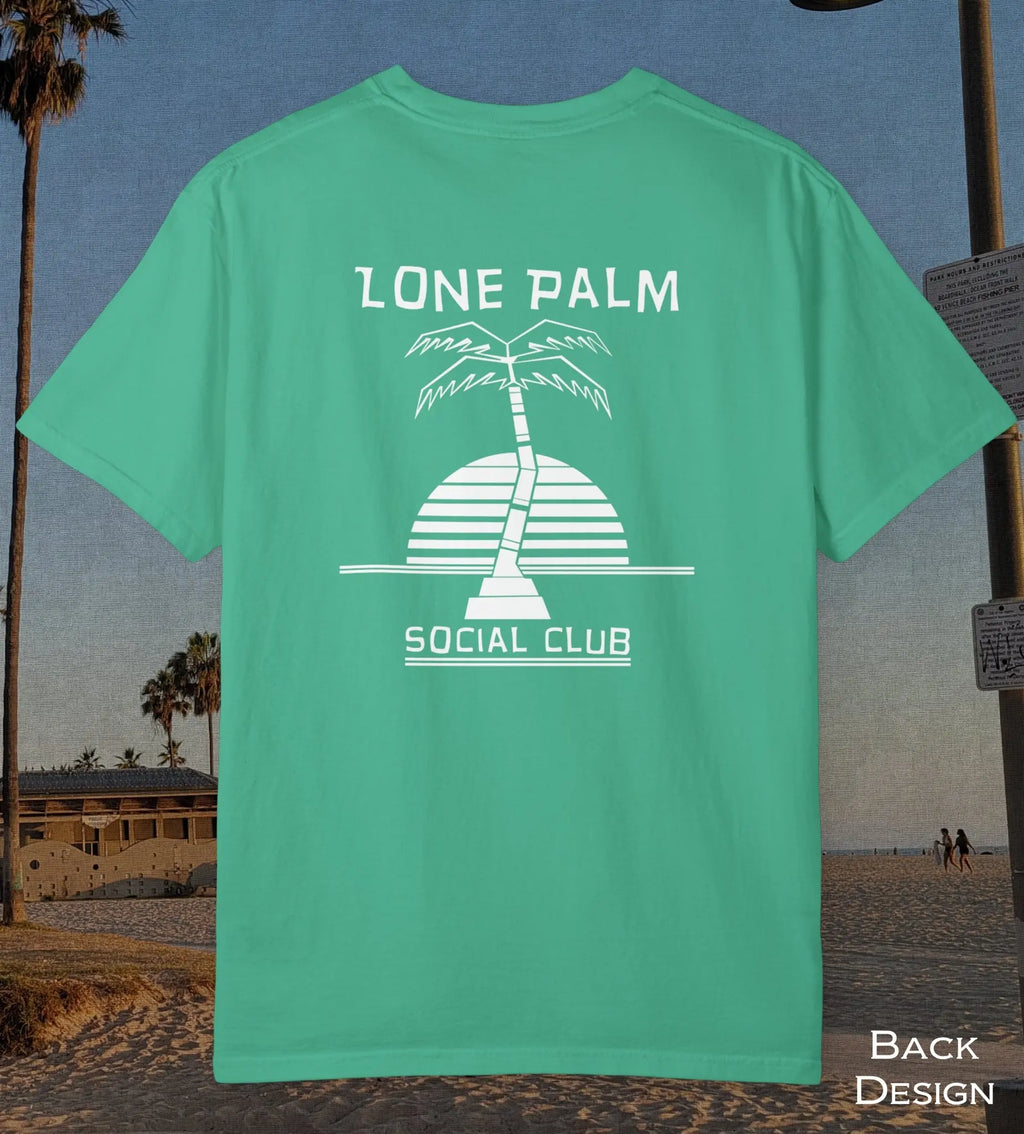 A mint green t-shirt shown from the back, featuring a white graphic of a palm tree and a striped sun with the text 'LONE PALM SOCIAL CLUB'. The t-shirt is displayed against a beach background.
