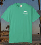 A mint green short-sleeve crewneck t-shirt with a small white graphic of a station wagon on the left chest, displayed against a beach background.