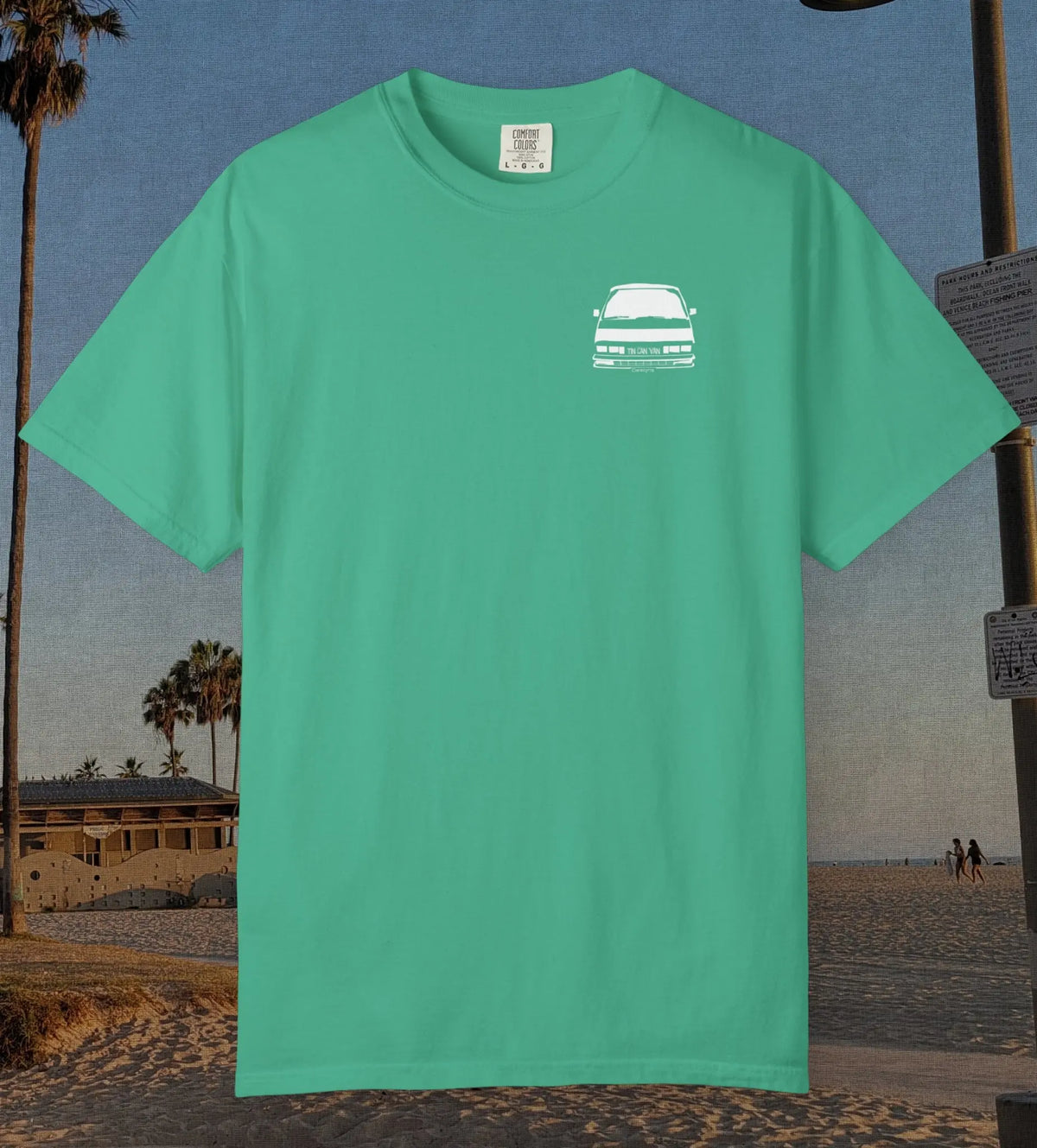A mint green short-sleeve crewneck t-shirt with a small white graphic of a station wagon on the left chest, displayed against a beach background.