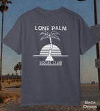 A navy t-shirt displayed on a beach background, showing the back design with a white graphic of a palm tree and a striped semi-circle sun. The text reads 'LONE PALM SOCIAL CLUB'.