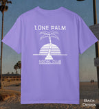 A violet purple t-shirt displayed on a beach background, showing the back design with a white graphic of a palm tree and a striped sun, with the text 'LONE PALM SOCIAL CLUB'.