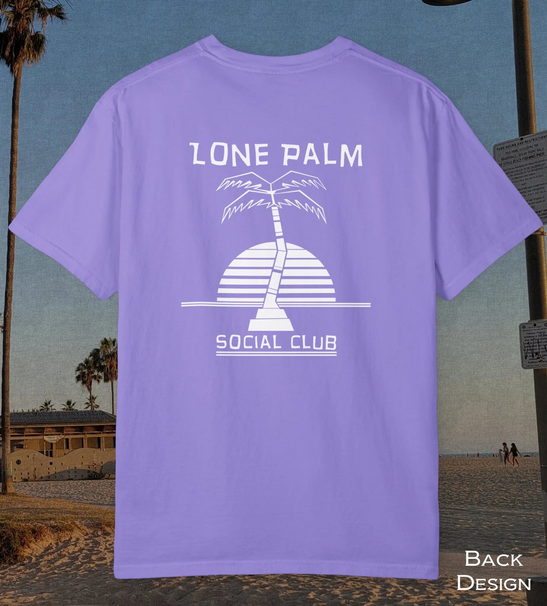 A violet purple t-shirt displayed on a beach background, showing the back design with a white graphic of a palm tree and a striped sun, with the text 'LONE PALM SOCIAL CLUB'.