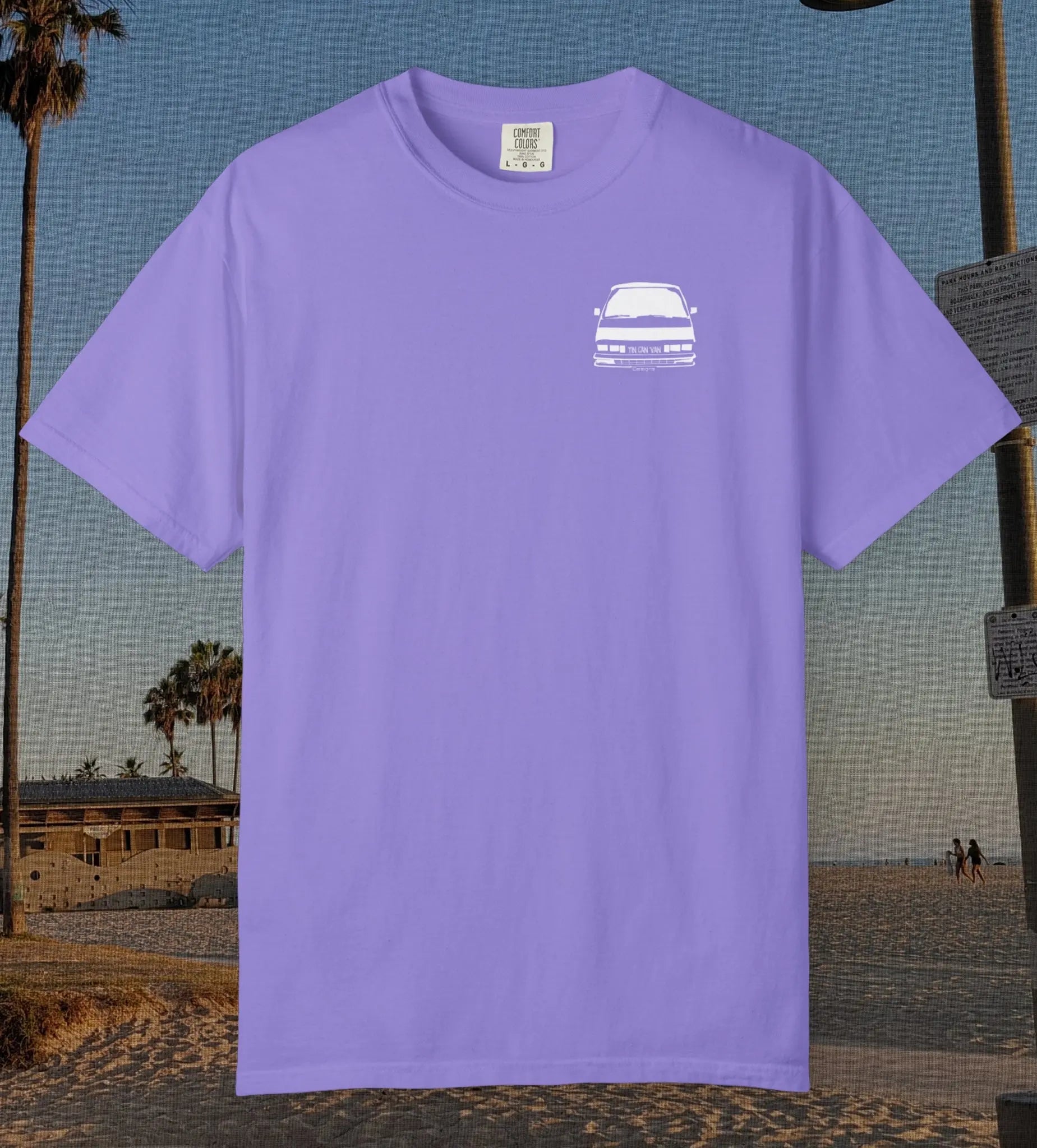 A violet short-sleeve crewneck t-shirt with a small white graphic of a classic car on the left chest, displayed against a beach background.