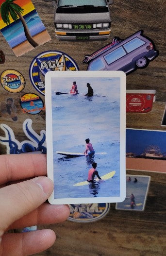 Hand holding a sticker with people on surfboards in water, surrounded by various stickers on a wooden surface.
