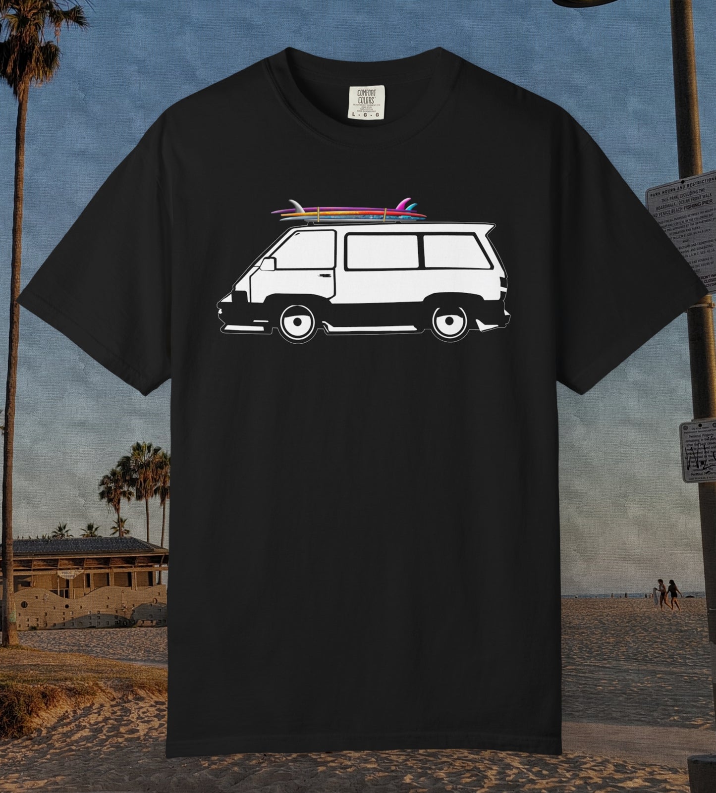 A black short-sleeve t-shirt featuring a white graphic of a classic camper van with pink and blue surfboards on its roof, displayed against a beach background.