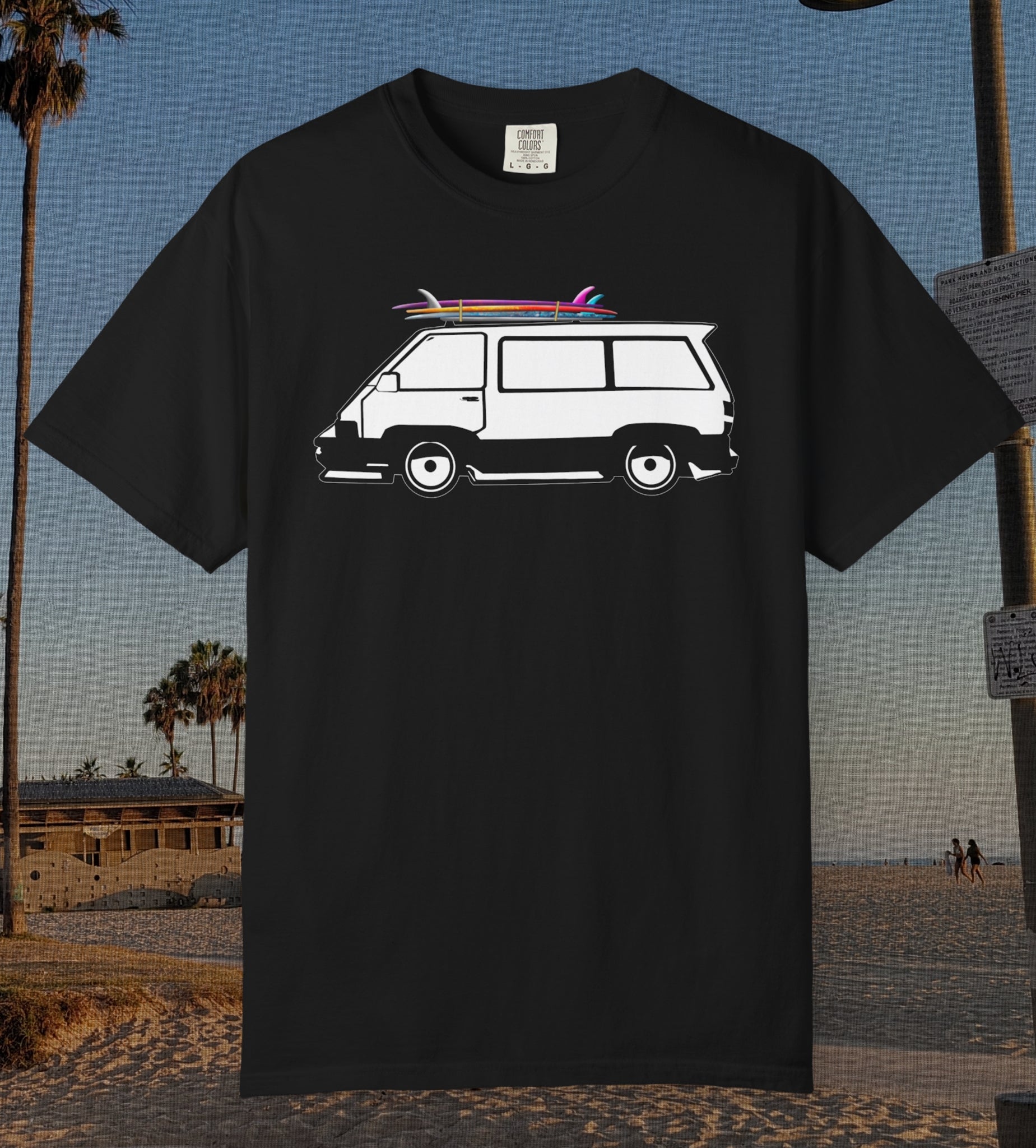 A black short-sleeve t-shirt featuring a white graphic of a classic camper van with pink and blue surfboards on its roof, displayed against a beach background.