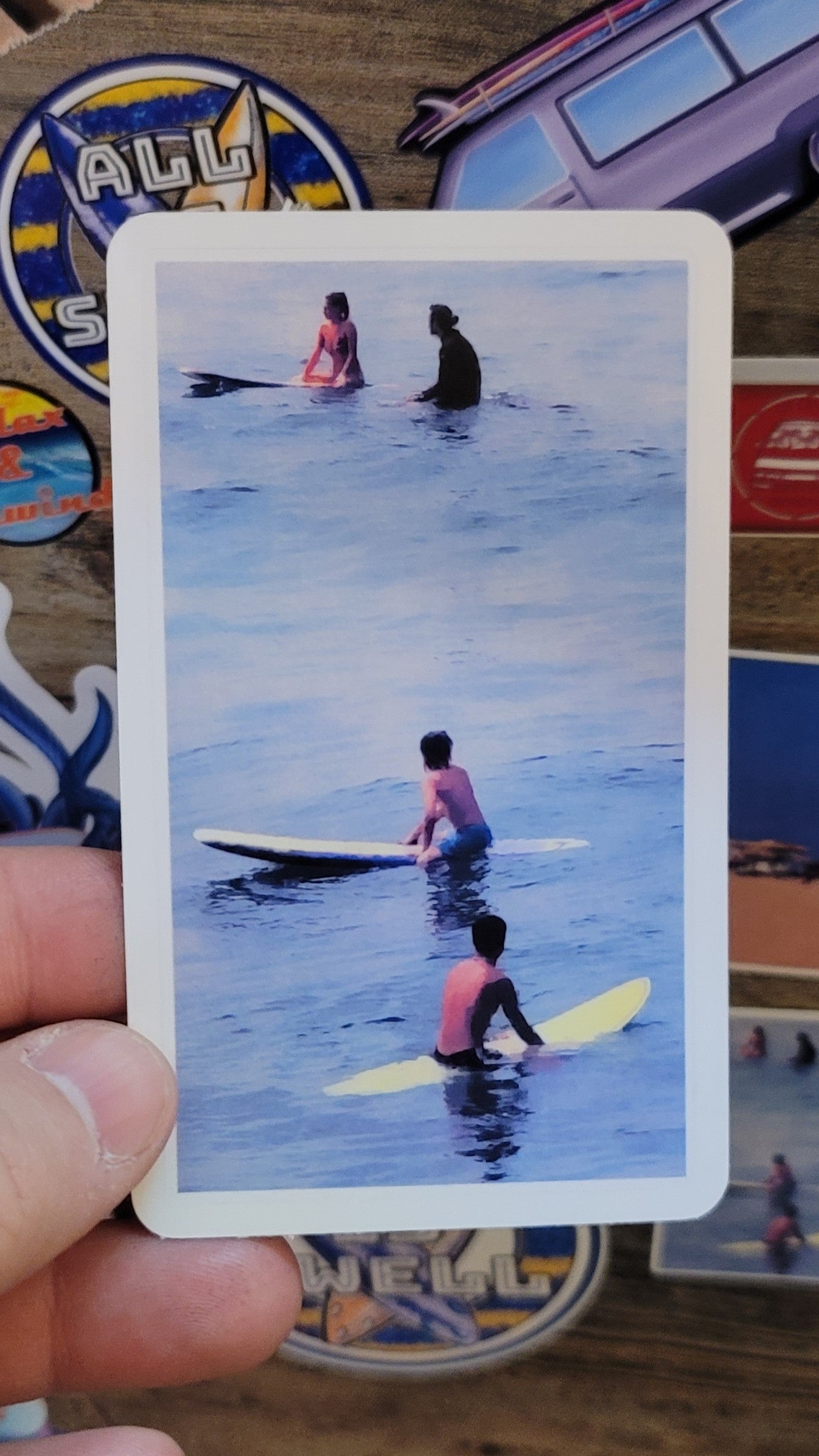 Sticker with surfers on surfboards held by a hand, aainst a wood background coverd by other stickers.