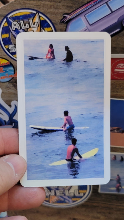 Sticker with surfers on surfboards held by a hand, aainst a wood background coverd by other stickers.
