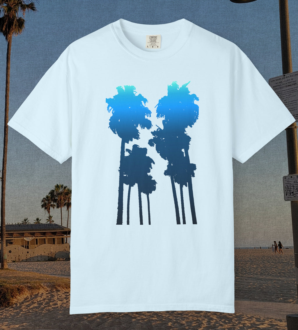 A chambray blue short-sleeve crewneck t-shirt featuring a graphic of two palm tree silhouettes filled with a blue-to-navy gradient, displayed against a beach background.