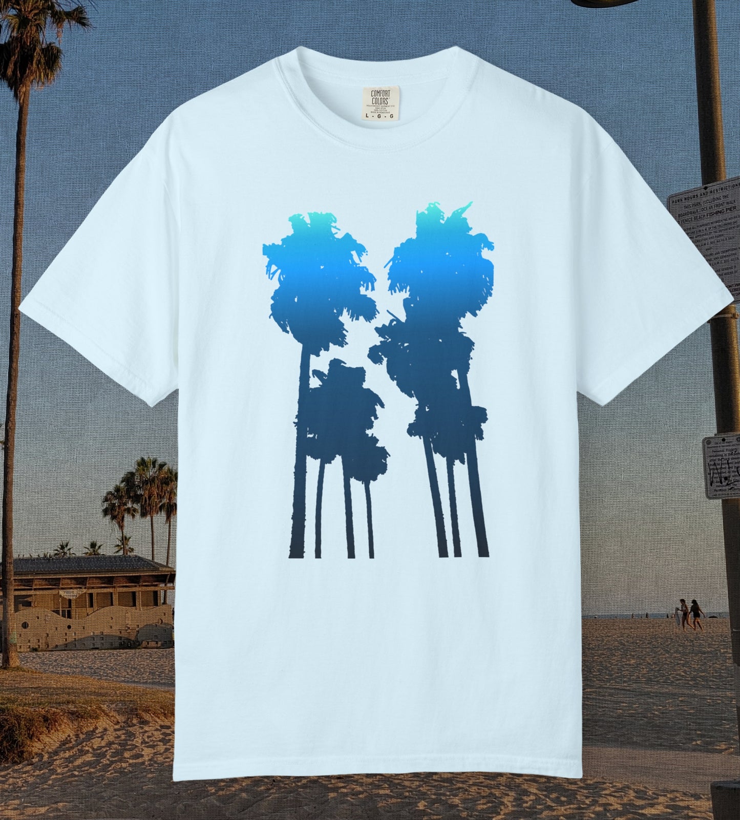 A chambray blue short-sleeve crewneck t-shirt featuring a graphic of two palm tree silhouettes filled with a blue-to-navy gradient, displayed against a beach background.