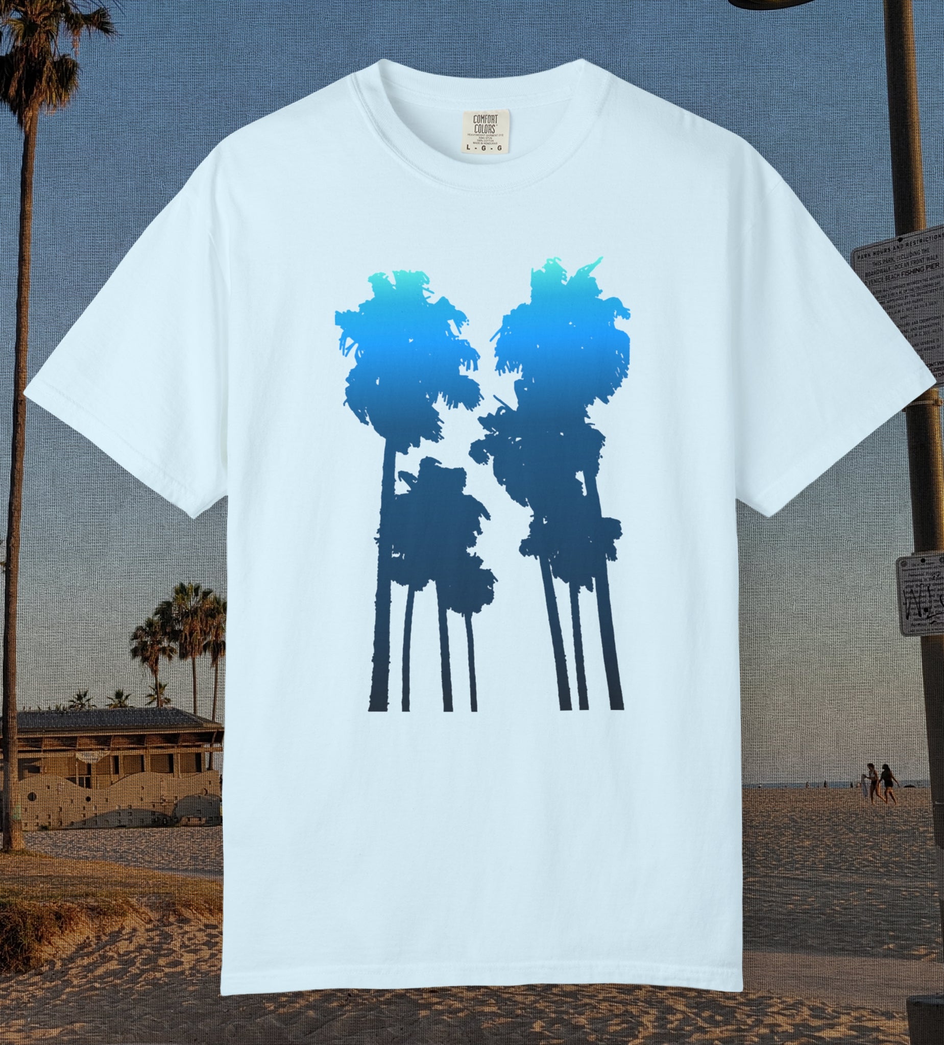 A chambray blue short-sleeve crewneck t-shirt featuring a graphic of two palm tree silhouettes filled with a blue-to-navy gradient, displayed against a beach background.