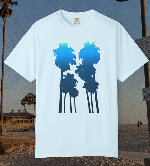 A chambray blue short-sleeve crewneck t-shirt featuring a graphic of two palm tree silhouettes filled with a blue-to-navy gradient, displayed against a beach background.