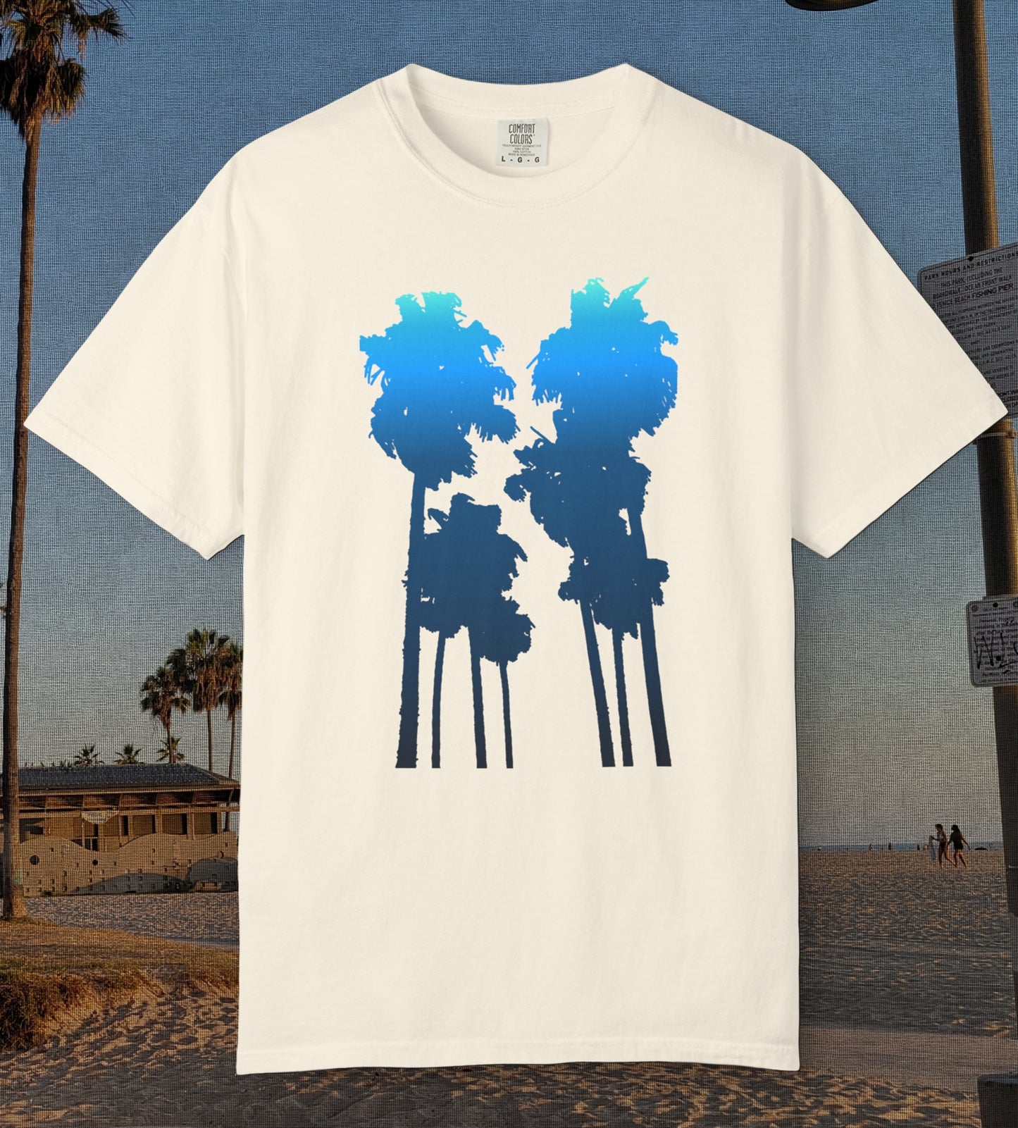 An ivory comfort colors short-sleeve t-shirt with a graphic of two palm tree silhouettes in light blue to navy blue gradient, displayed against a beach background.