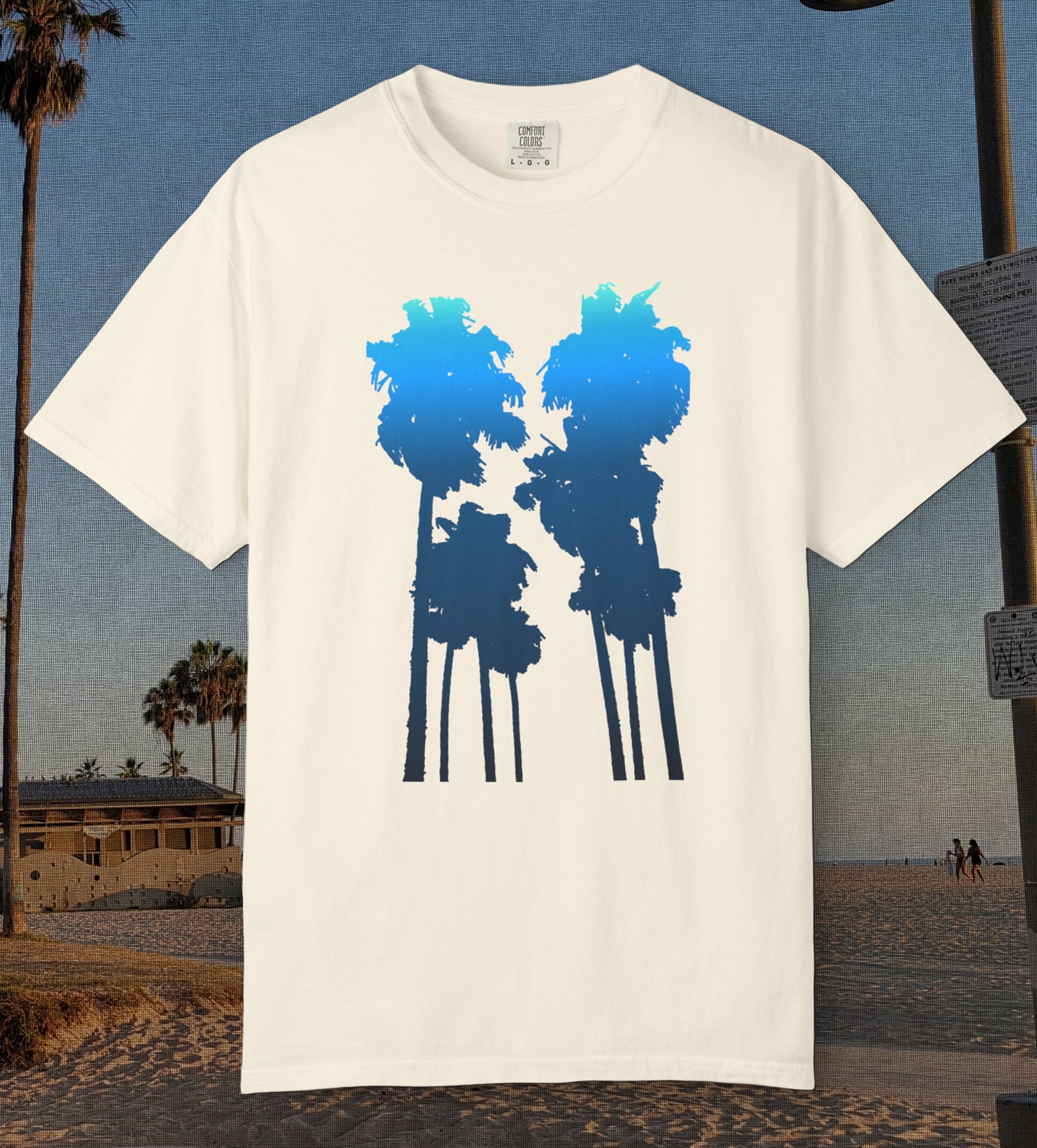 An ivory comfort colors short-sleeve t-shirt with a graphic of two palm tree silhouettes in light blue to navy blue gradient, displayed against a beach background.
