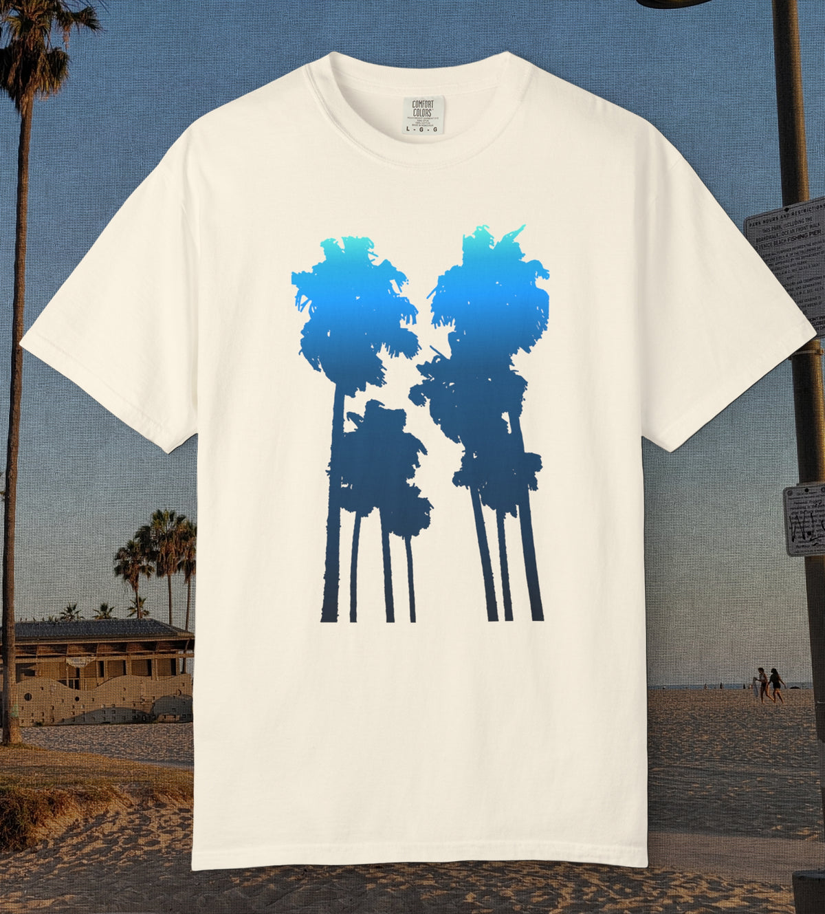 An ivory comfort colors short-sleeve t-shirt with a graphic of two palm tree silhouettes in light blue to navy blue gradient, displayed against a beach background.