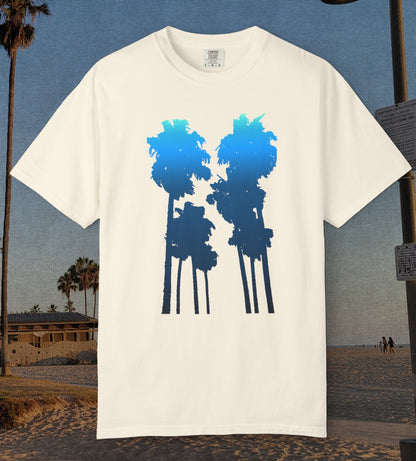 An ivory comfort colors short-sleeve t-shirt with a graphic of two palm tree silhouettes in light blue to navy blue gradient, displayed against a beach background.